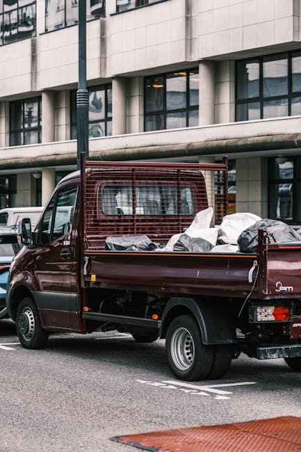 A small dark red flatbed truck is parked on an urban street in front of a modern building with large rectangular windows and a stone facade. The truck's cargo bed is filled with assorted waste items,