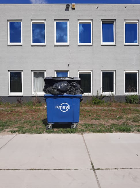 A small dark red flatbed truck is parked on an urban street in front of a modern building with large rectangular windows and a stone facade. The truck's cargo bed is filled with assorted waste items,