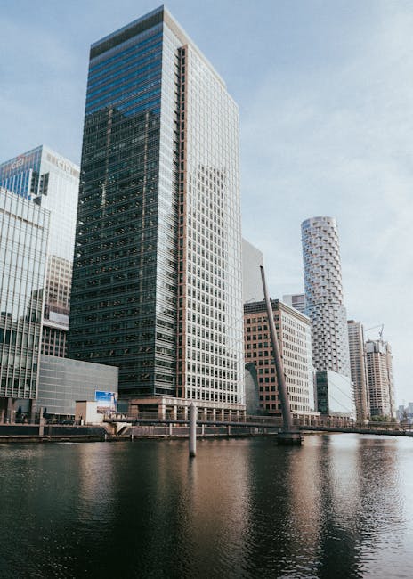 The image displays a modern cityscape with a prominent tall skyscraper featuring a glass façade reflecting nearby buildings, situated along a river or canal. Adjacent structures include other high-ri