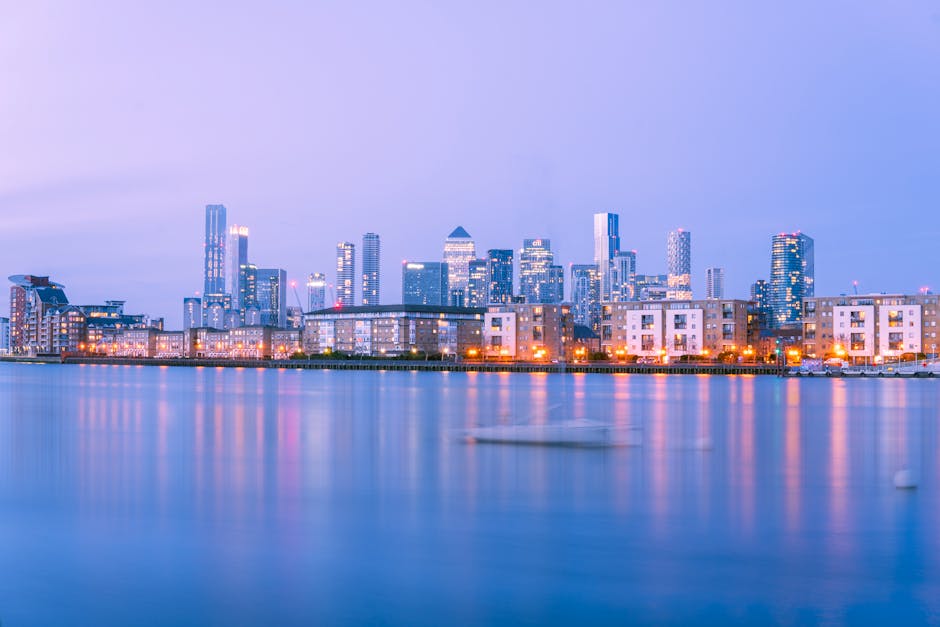 The image displays a modern cityscape with a prominent tall skyscraper featuring a glass façade reflecting nearby buildings, situated along a river or canal. Adjacent structures include other high-ri